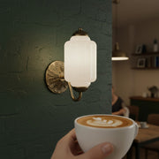 A warm vintage milk glass sconce glowing on a green wall in a cozy cafe, with a latte in the foreground.